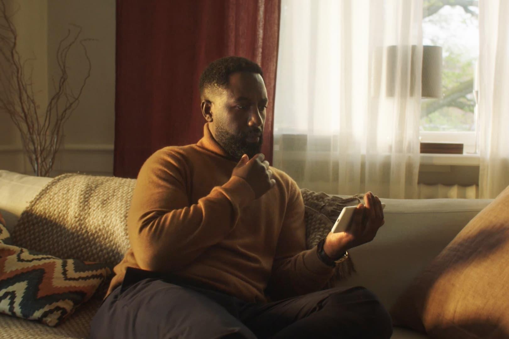 Person som sitter i en soffa och ser fundersam ut. Personen håller i en mobil