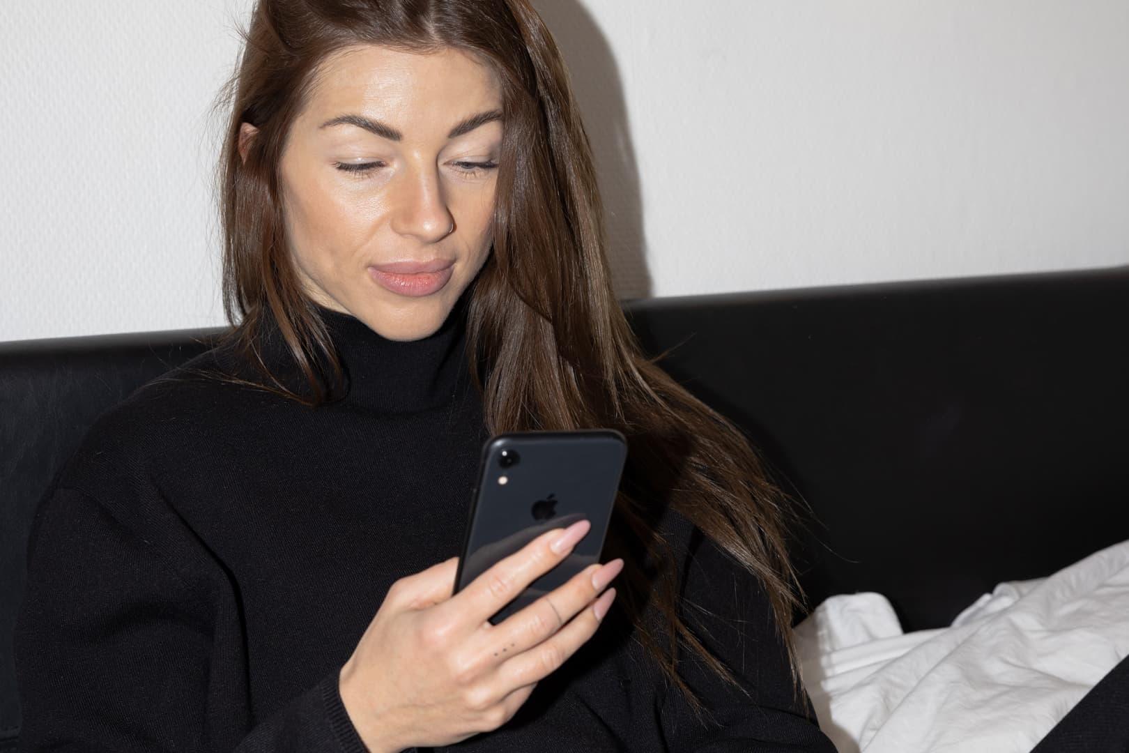 En kvinna med långt brunt hår som sitter i en soffa hemma hos sig och tittar på sin mobil.